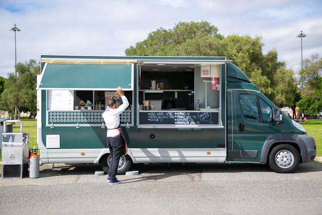 Green food truck with an open service window serving a customer at a park.
