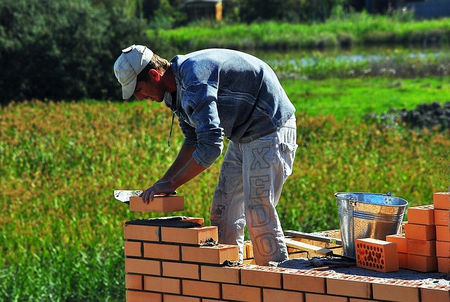 Construction worker lays bricks to build a low wall outdoors, with a metal bucket nearby.